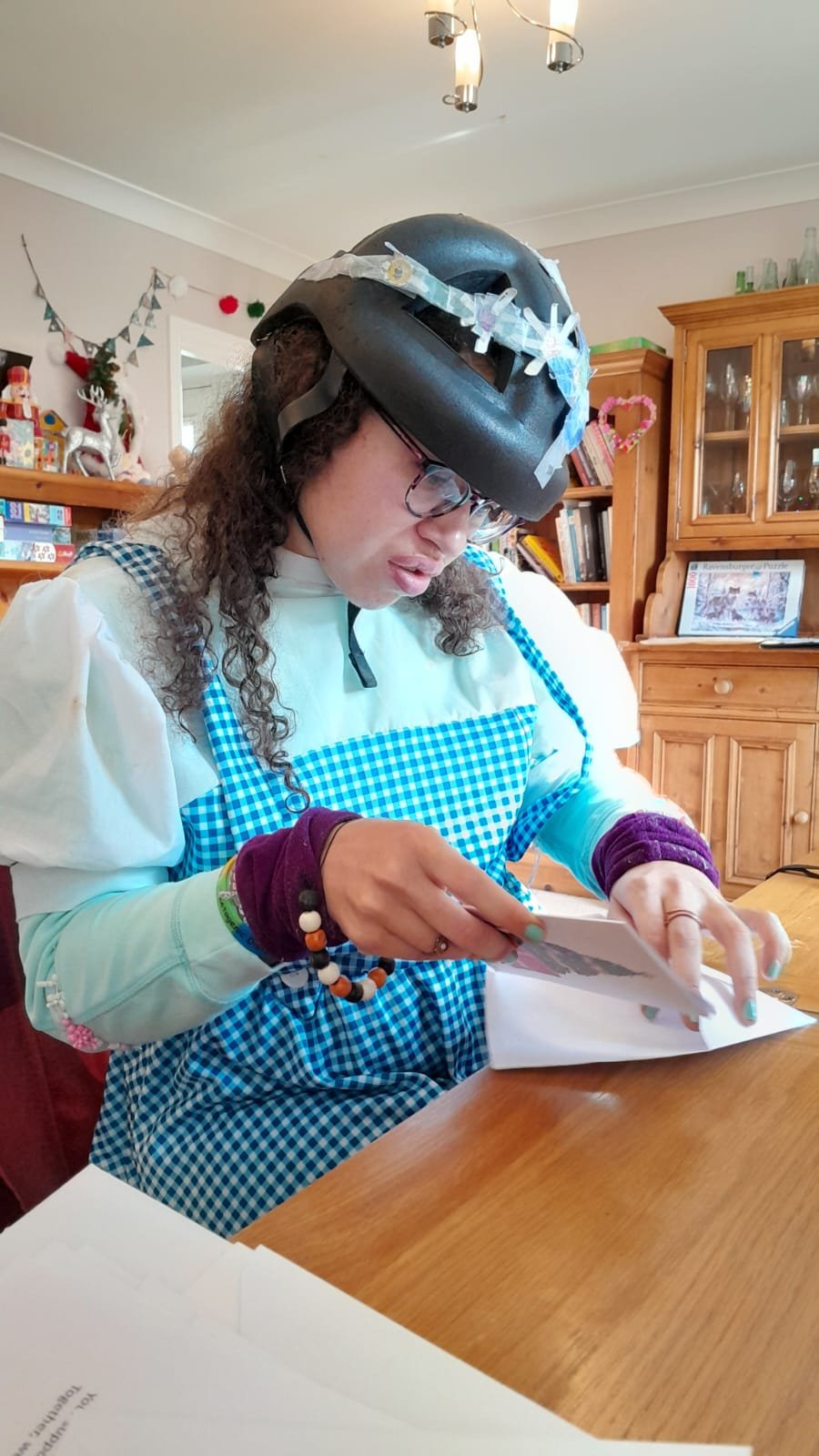 A woman wearing a helmet with tape on it, glasses, a white shirt, a blue gingham apron, and purple wristbands, sitting at a wooden table, looking at a photograph or card.