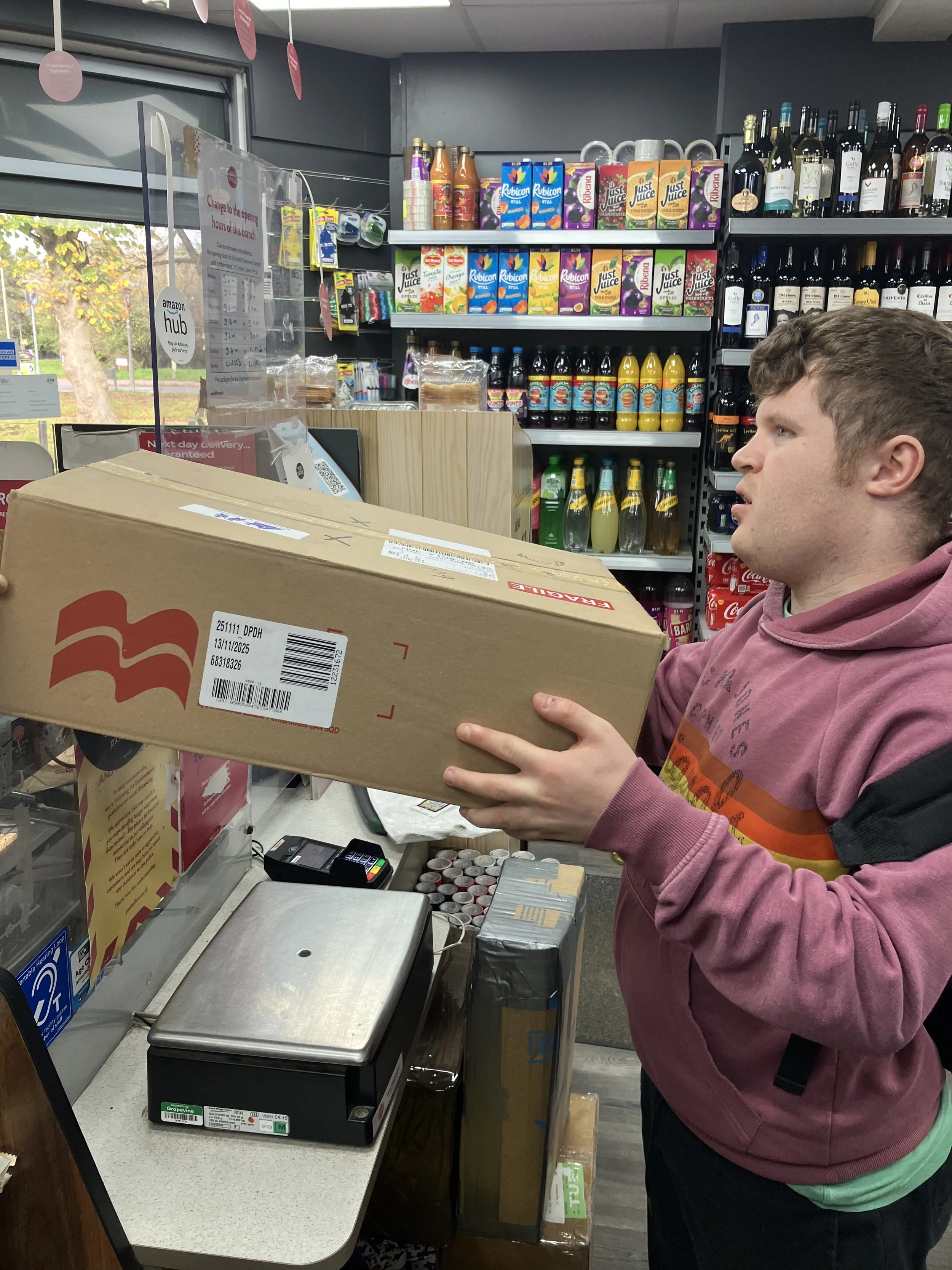 A young man in a pink hoodie at a checkout counter holding a large cardboard box in a grocery store. Shelves of beverages and snacks are visible in the background.