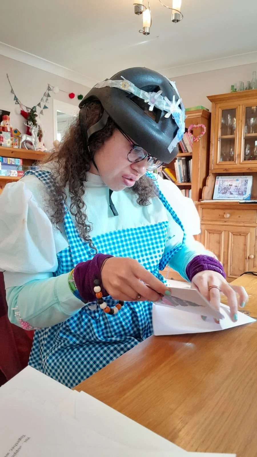A woman wearing a black bike helmet decorated with paper clips, a blue checkered apron, and purple wristbands sits at a wooden table reading a piece of paper inside a room with bookshelves and festive decorations.