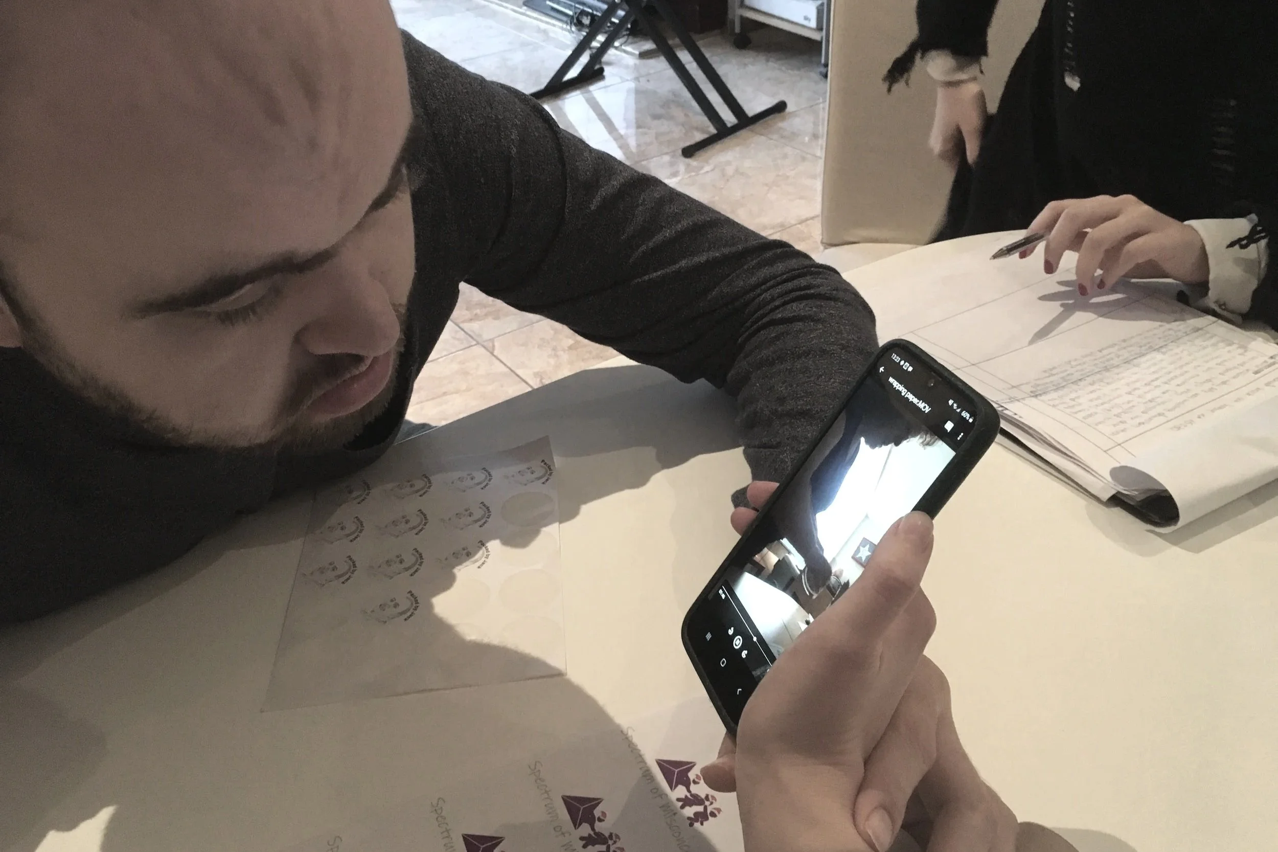 A man is lying on a table, looking at his smartphone, with stickers of a leaf and an eye on the table. In the background, a person is writing on a notebook.