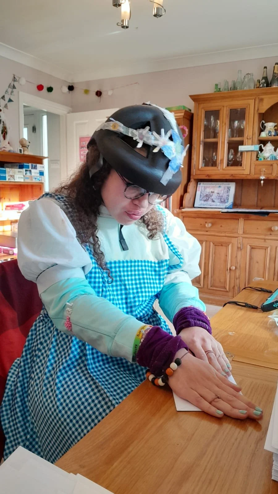 A woman wearing a black helmet decorated with ribbons, glasses, and a blue and white gingham apron sitting at a wooden table, focusing on an activity with her hands. The background shows a cozy room with wooden furniture and decorations.