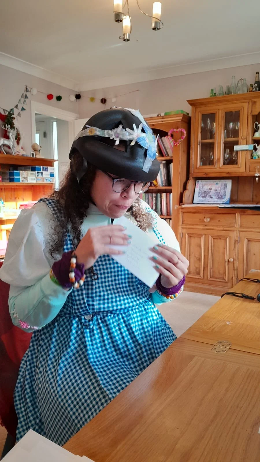Woman wearing a black helmet decorated with paper flowers and ribbons, reading a handwritten card, sitting in a cozy wooden-furnished room with holiday decorations.