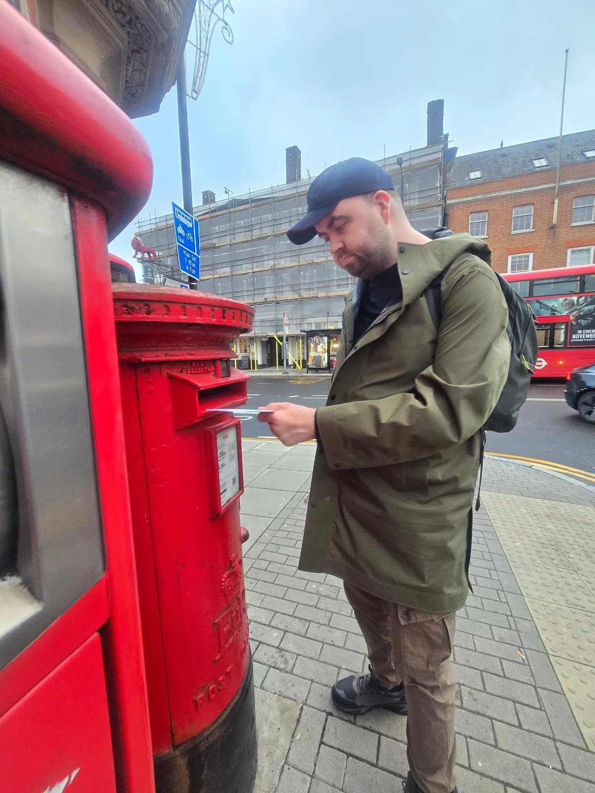 A man in a green jacket and black cap using a red mailbox on a city sidewalk.