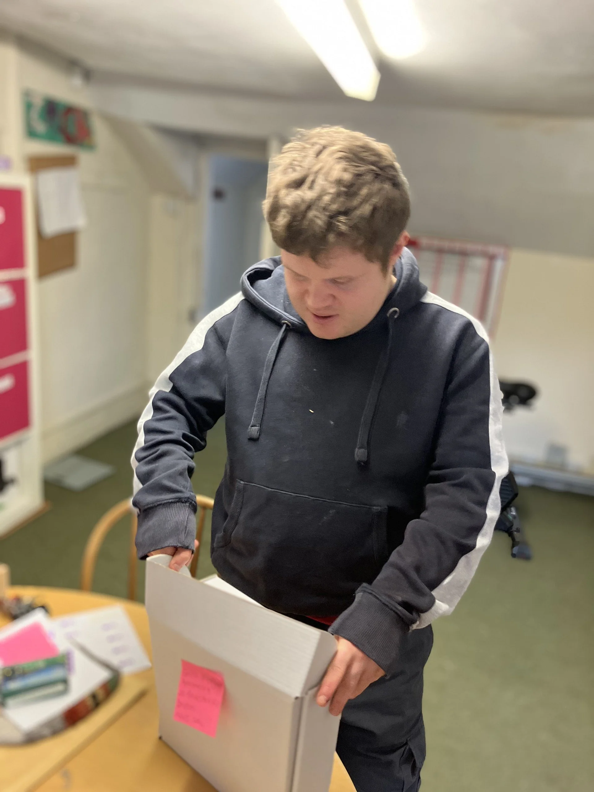 A teenage boy with short brown curly hair, wearing a black hoodie with white stripes on the sleeves, standing at a table and opening a white cardboard box with a pink sticky note on it.