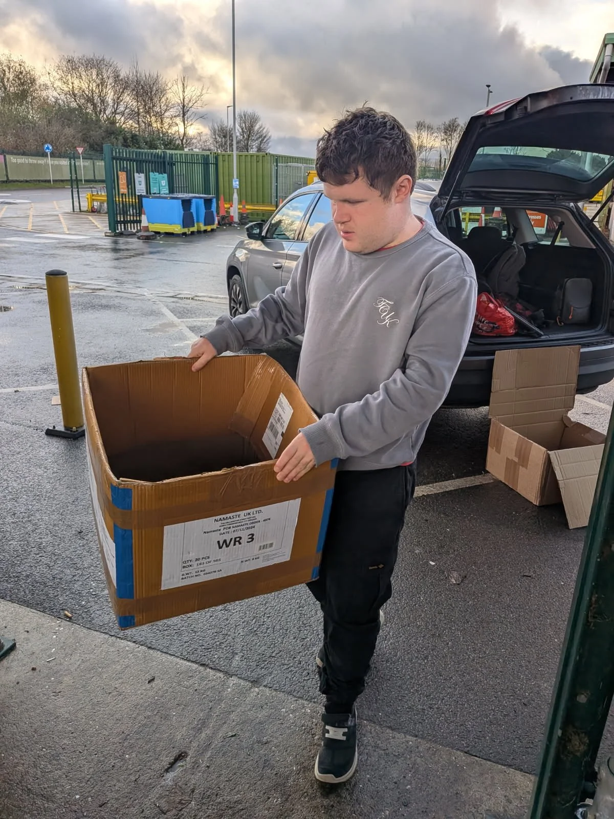 A young man is standing outside near a car, holding an empty cardboard box. The car's trunk is open behind him, and there are other cardboard boxes nearby. The parking lot appears wet, and there are trees, a fence, and a cloudy sky in the background.