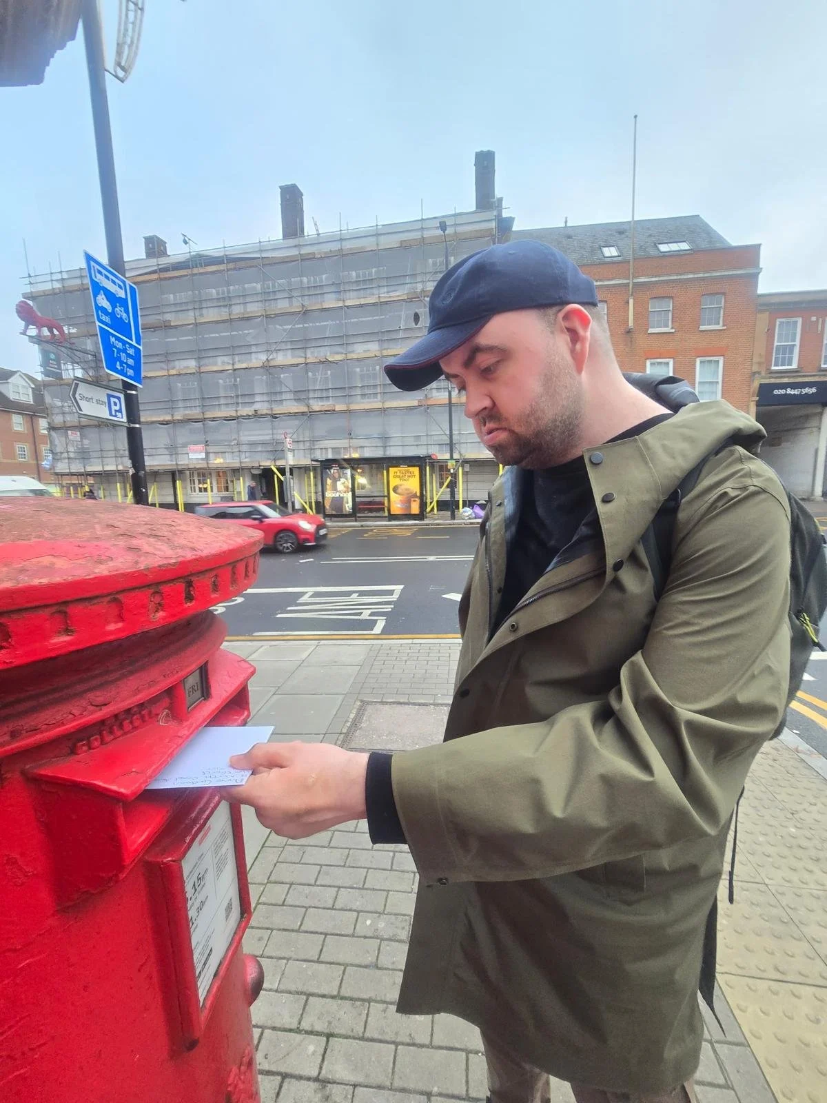 Man in a green jacket and black cap dropping a letter into a red mailbox on a city sidewalk.
