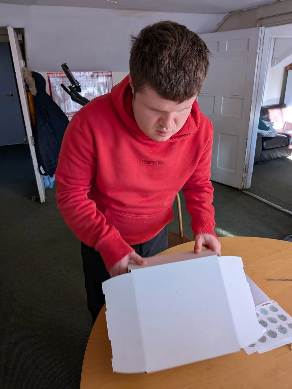 A young man in a red hoodie looking inside an open pizza box on a wooden table.