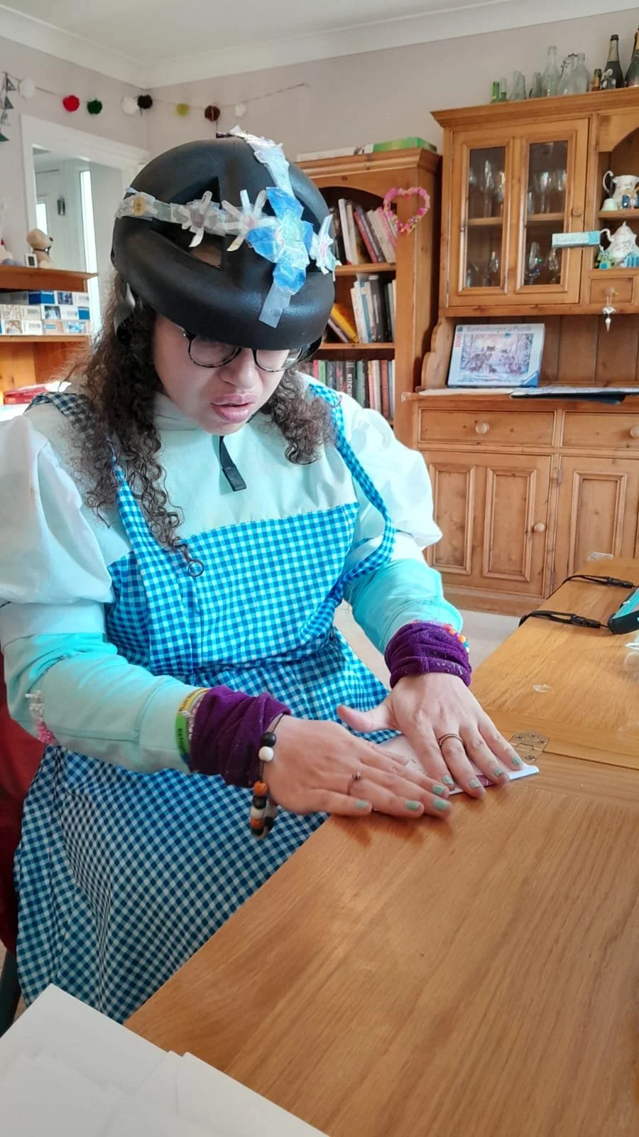 A person wearing a decorative helmet, glasses, a white and blue checkered apron, and purple wristbands, is working on a paper at a wooden table in a cozy room with wooden cabinets and bookshelves.
