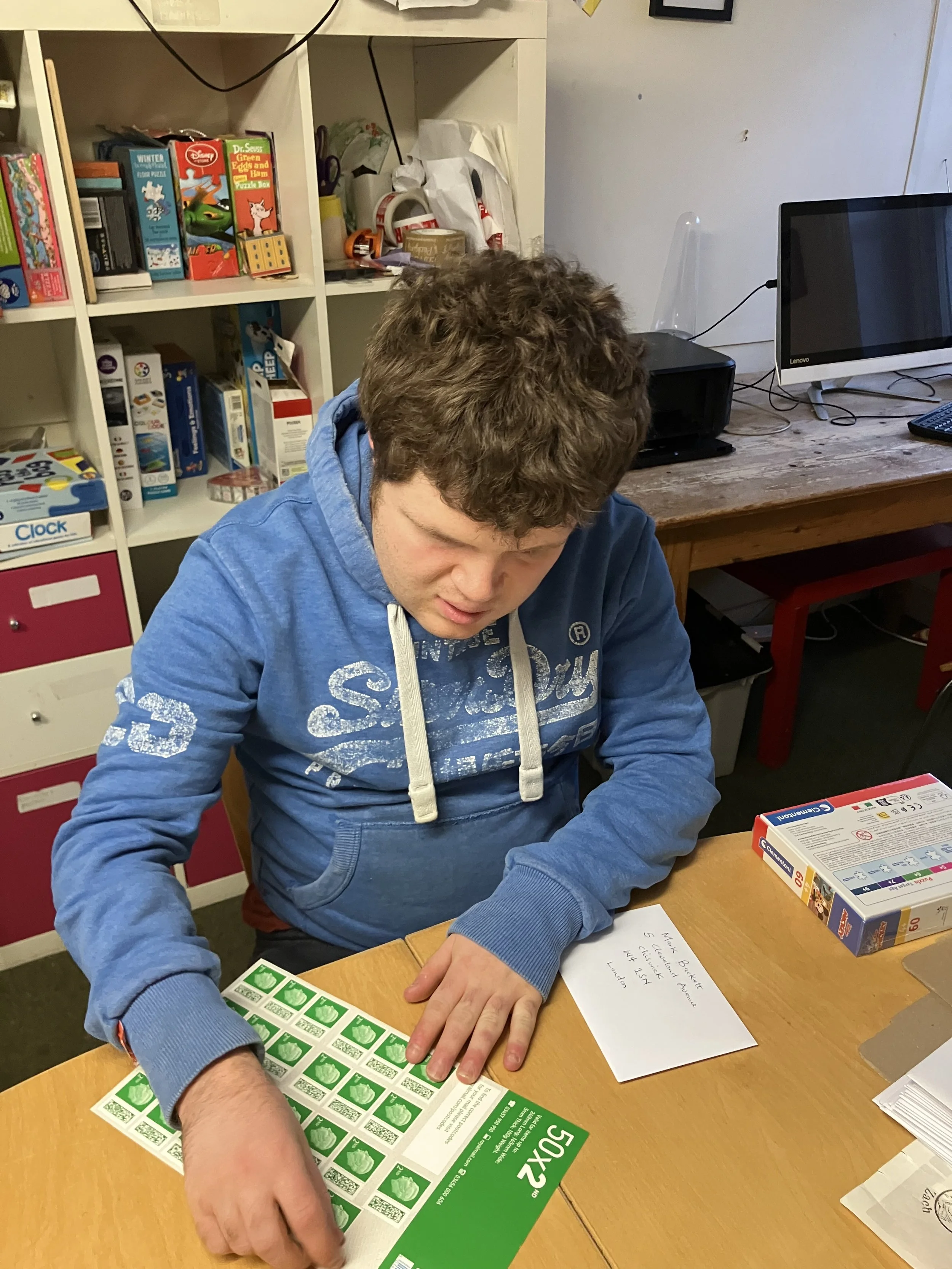 A young man in a blue hoodie is sitting at a wooden table, counting green food coupons. There is a note on the table and a box of board game pieces nearby. In the background, there are shelves with board games and a computer on a desk.