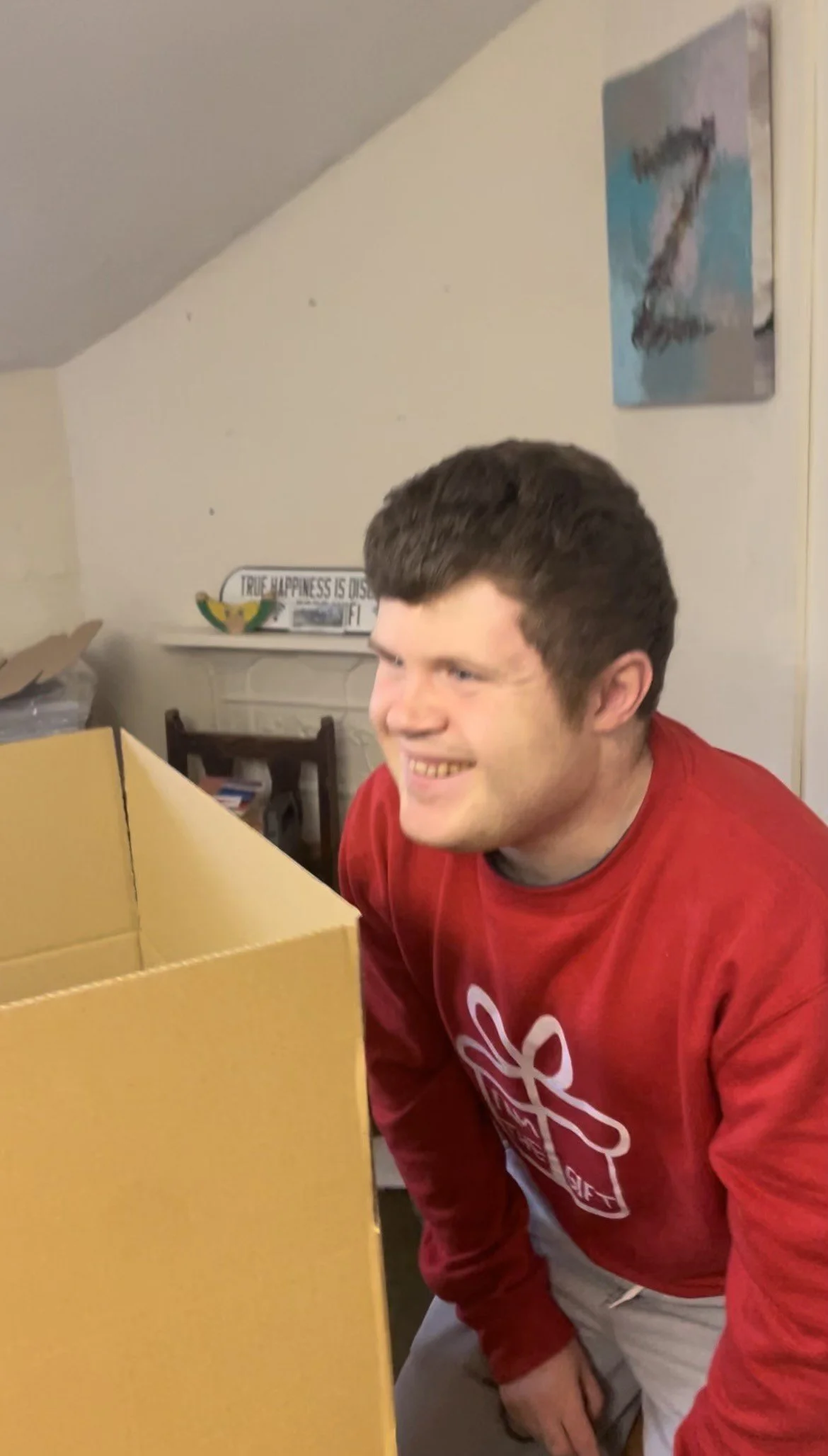 A young man with short brown hair is smiling while looking into a large cardboard box. He is wearing a red sweatshirt with a white gift box graphic and gray shorts. The setting appears to be a room with a white wall, some art, a sign, and furniture i