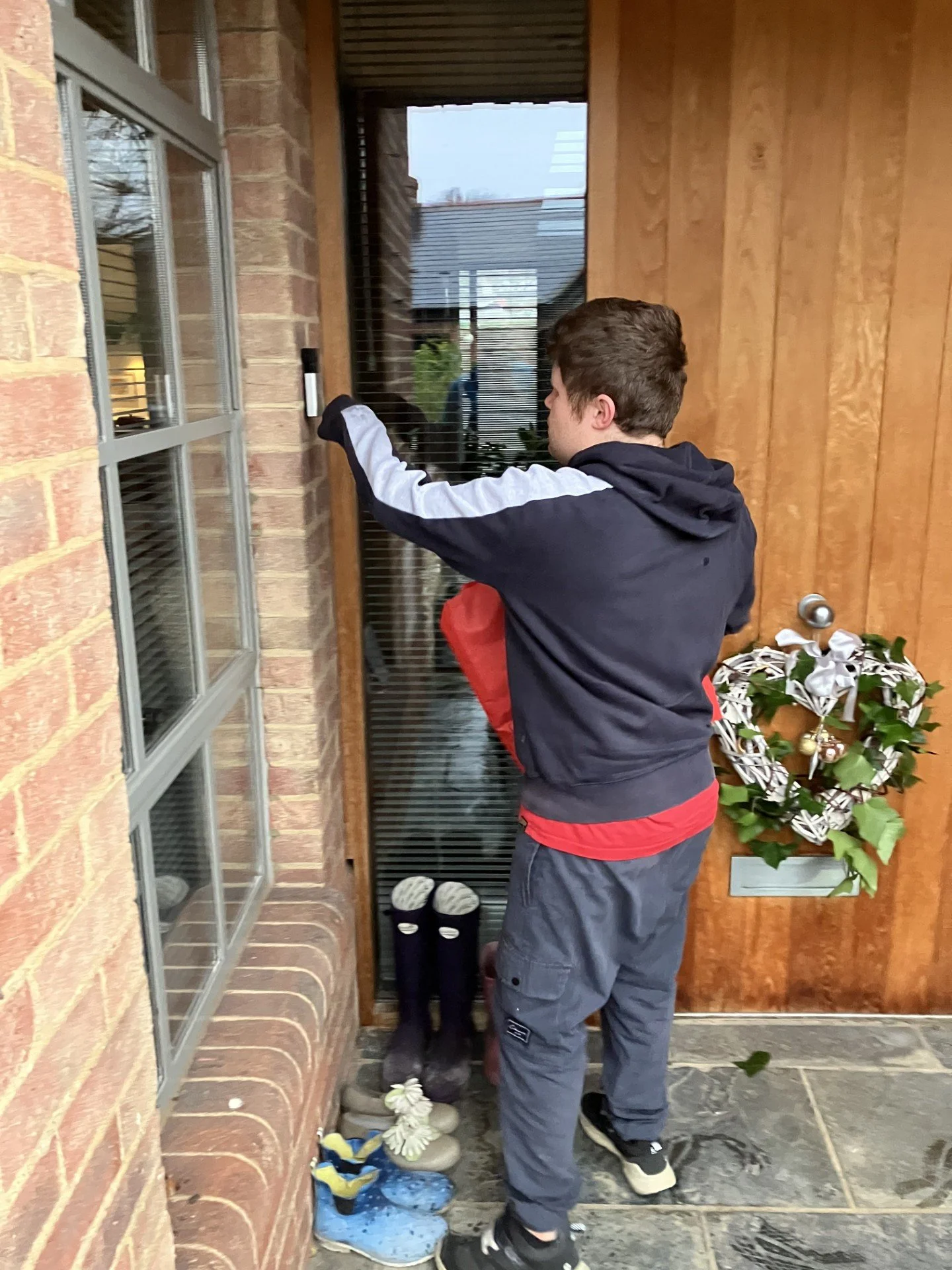 A young boy in a black and red jacket and gray pants standing at a front door, ringing the doorbell. There are shoes and boots on the floor near the door and a holiday wreath hanging on the wooden door.