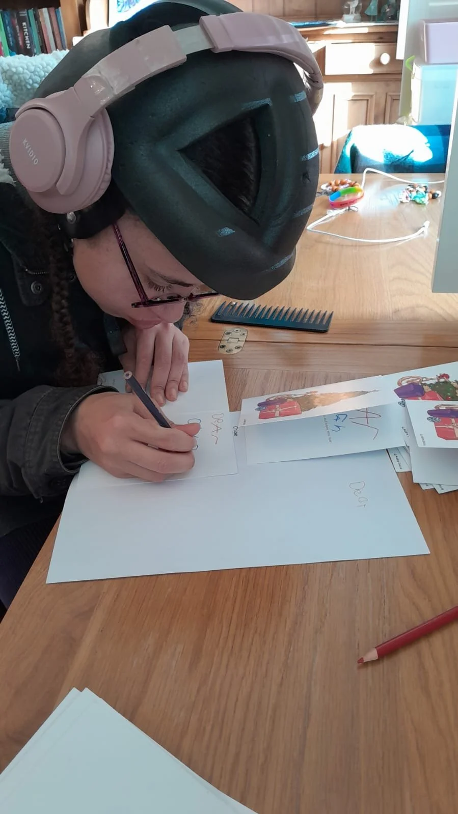 A woman wearing a black bike helmet and pink headphones is sitting at a wooden table, writing on a white sheet of paper with a black pen. There are birthday and holiday cards, a red pencil, and miscellaneous items on the table, with sunlight streamin