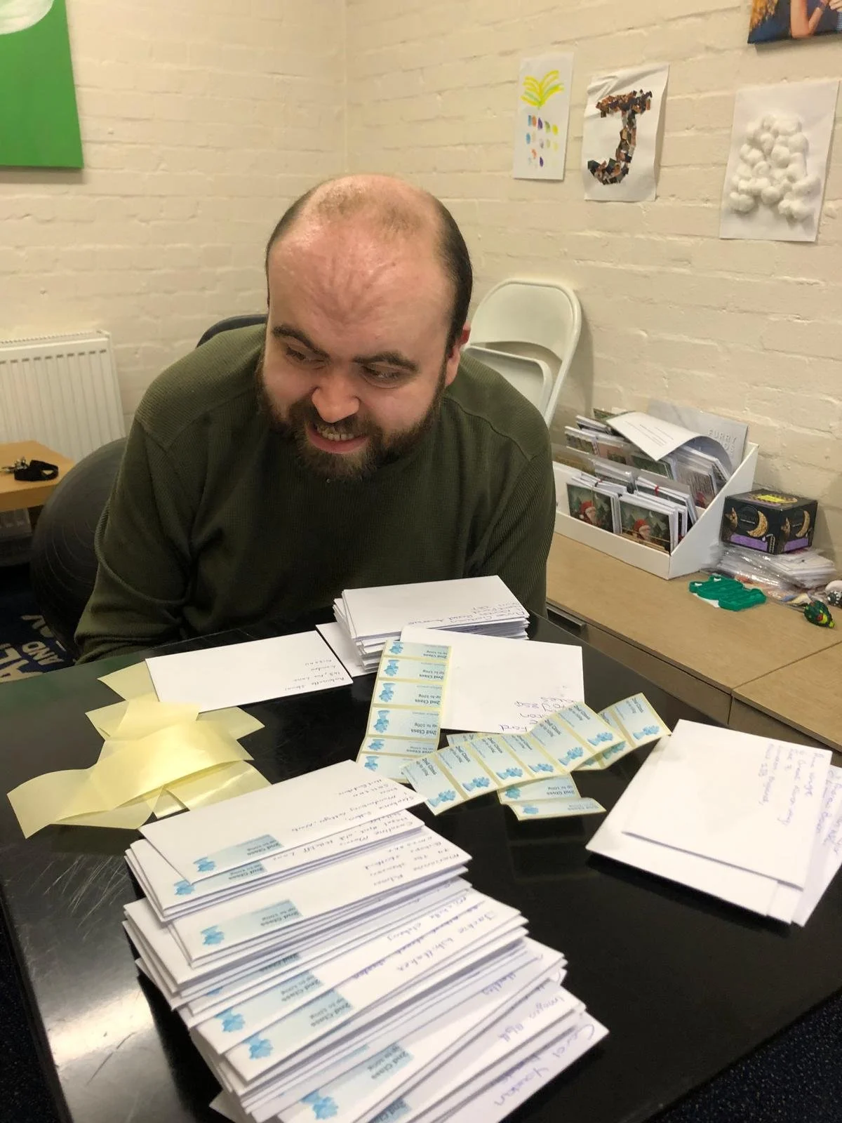 A man sitting at a table with piles of paper and stamps, smiling, with artwork on the brick wall behind him.