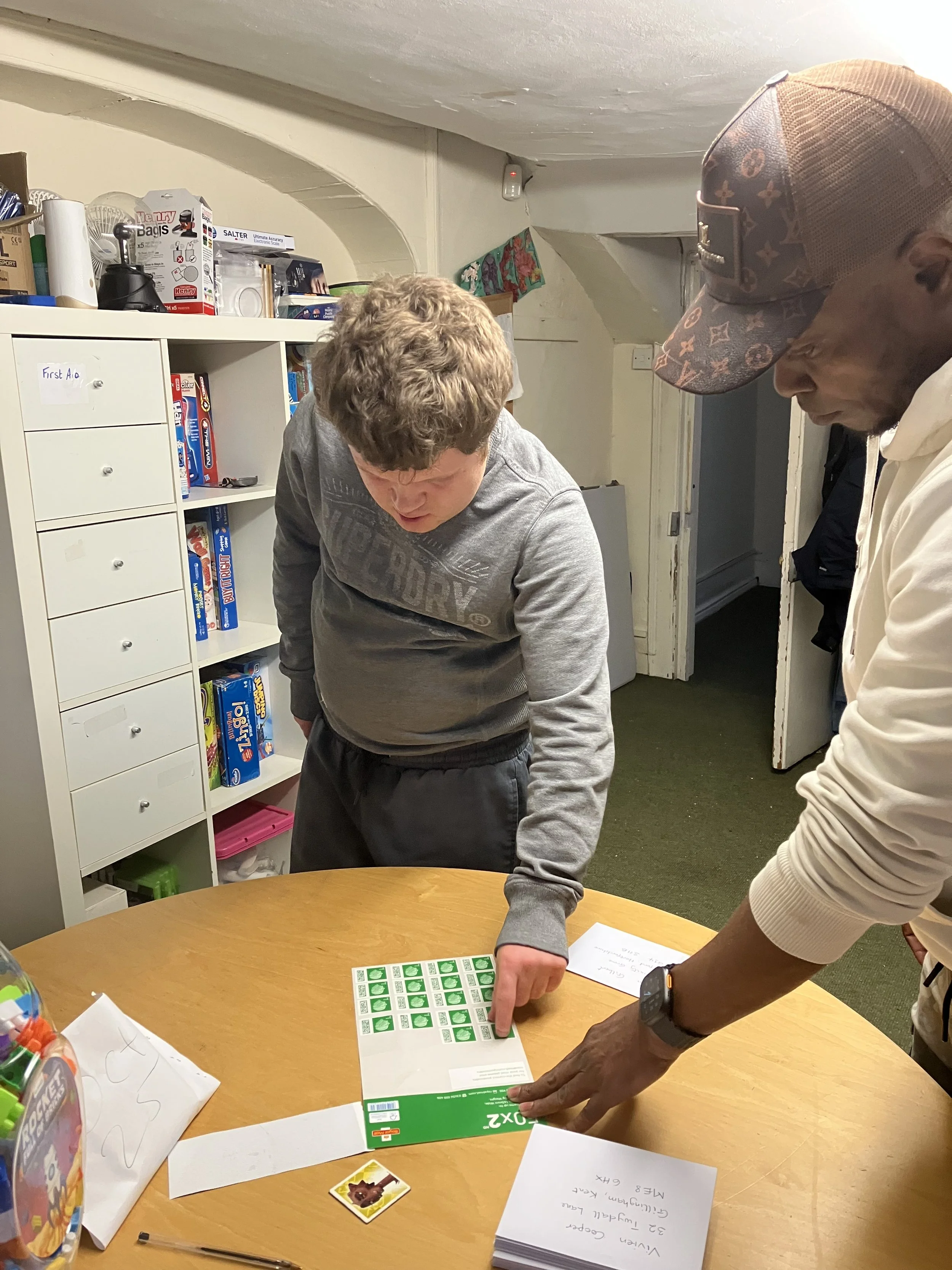 Two men playing bingo at a round table in a cozy room with white shelving and storage behind them. One man is pointing at his bingo card while the other observes.