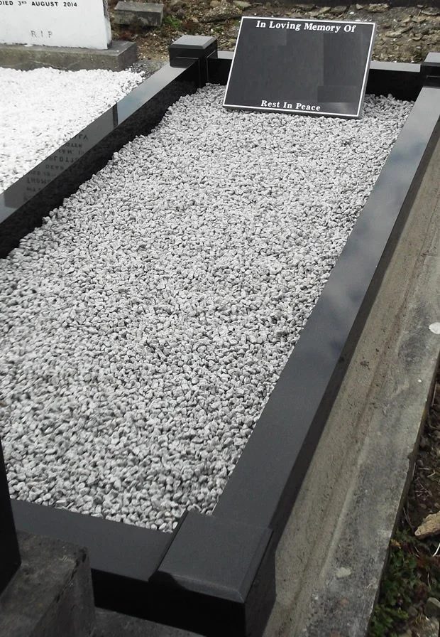 Grave at a cemetery with a black border, filled with small gray stones. A black and white memorial plaque on top reads 'In Loving Memory Of,' but the name and details are not visible.