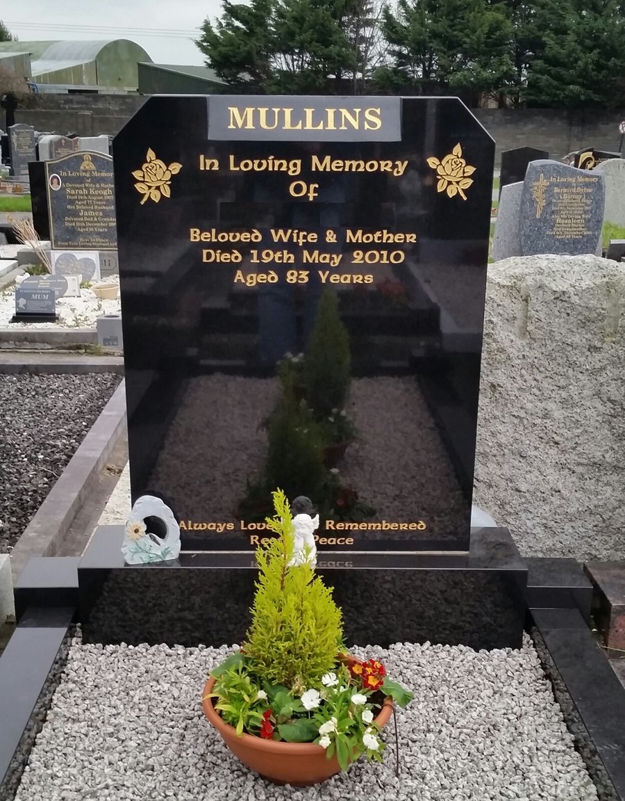 Black granite headstone with gold lettering in a cemetery. It reads 'MULLINS In Loving Memory Of Beloved Wife & Mother Died 19th May 2010 Aged 83 Years'. There are decorative roses on top. A small flower pot with colorful flowers and a cherub figurine are in front of the headstone.
