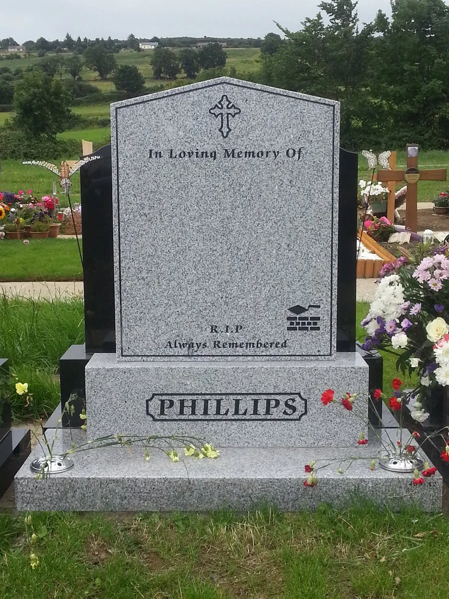 Gray granite headstone in a cemetery with flowers and grass around it. The headstone has a cross at the top and the words "In Loving Memory Of" and "R.I.P Always Remembered," with the surname "PHILLIPS" engraved at the bottom. There are decorative pinwheels and floral arrangements nearby.