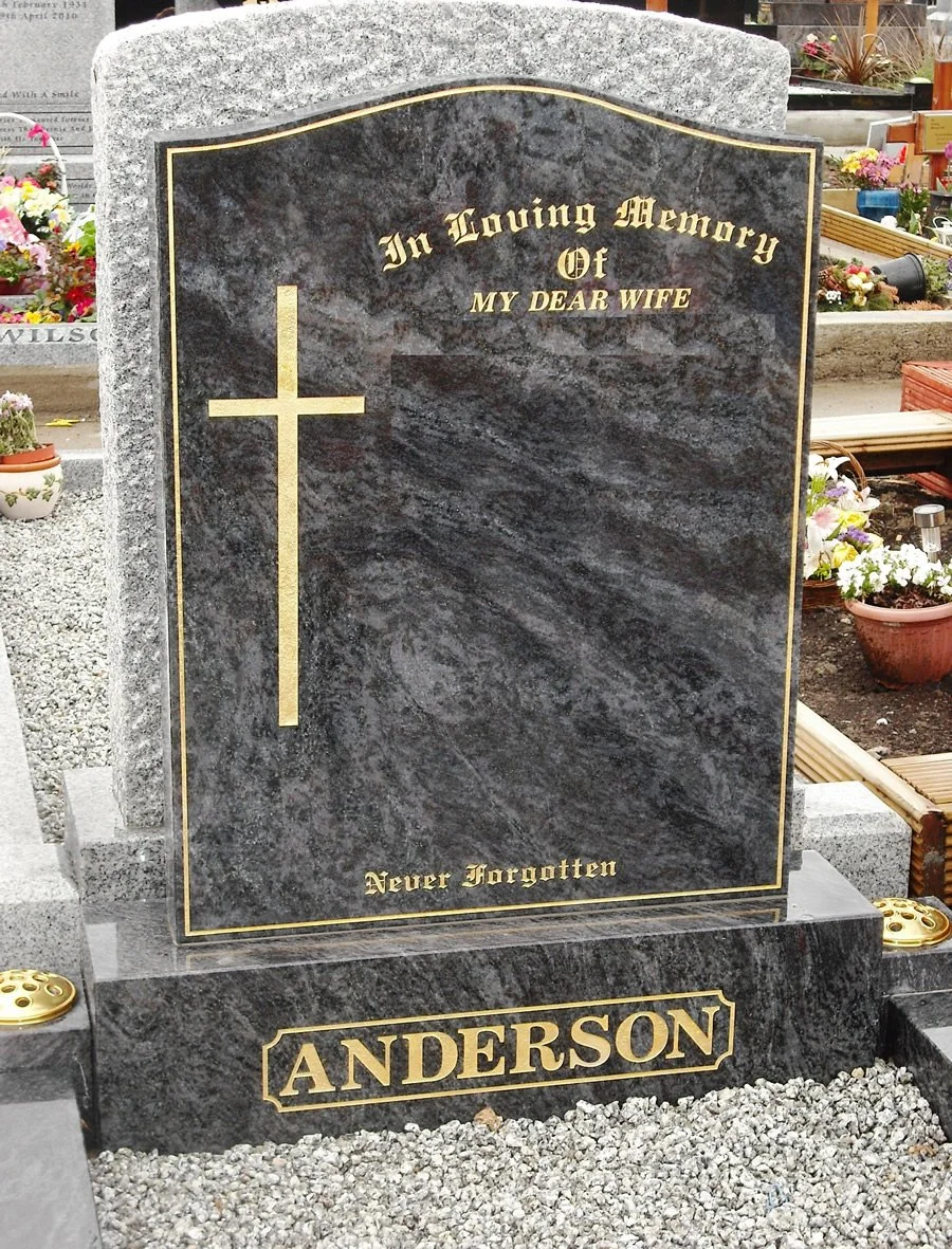Black and gray marble headstone with gold engraved text reading "In Loving Memory Of My Dear Wife" and "Never Forgotten," with a gold cross on the left side, located in a cemetery with flowers around.