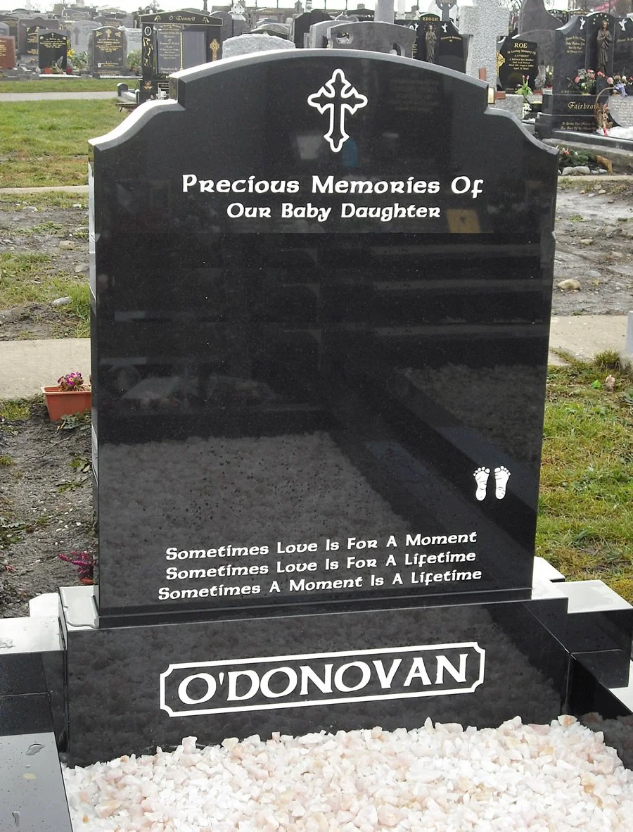 Black granite headstone with religious cross symbol at the top, and inscriptions dedicated to a baby daughter, with a pair of small footprints engraved and a quote about love. The headstone displays the name O'DONOVAN at the bottom, surrounded by white gravel at the base.