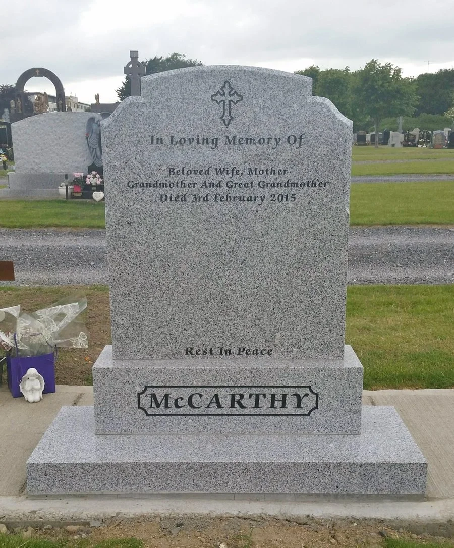 A gray granite tombstone with a cross on top, inscribed in memory of a beloved wife, mother, grandmother, and great grandmother who died on February 3, 2015. The surname 'McCARTHY' is engraved at the bottom, and the phrase 'Rest In Peace' is also inscribed.