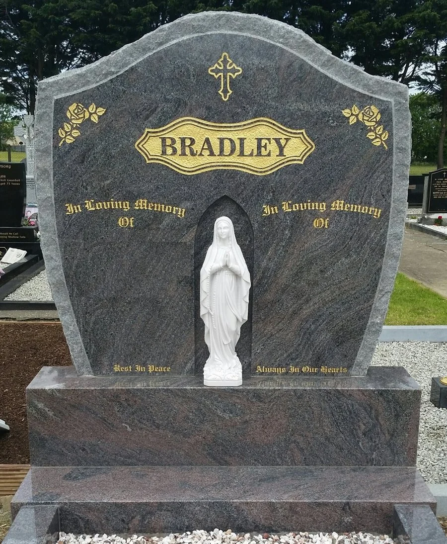 A funeral headstone with a carved statue of the Virgin Mary in the center. The headstone is made of dark stone with gold lettering and decorative roses. It has the name 'Bradley' at the top, a cross above, and inscriptions that read 'In Loving Memory Of', 'Rest In Peace', and 'Always In Our Hearts' on it.