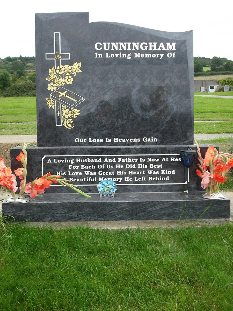 Gravestonein a cemetery with flowers and a small decorative globe. The gravestone features a Christian cross, flowers, and a Bible. It bears the inscription: "Cunnigham In Loving Memory Of. Our Loss Is Heavens Gain. A Loving Husband And Father Is Now At Rest For Each Of Us He Did His Best. His Love Was Great His Heart Was Kind. Beautiful Memory He Left Behind." The background includes green grass, open fields, and no other tombstones visible.
