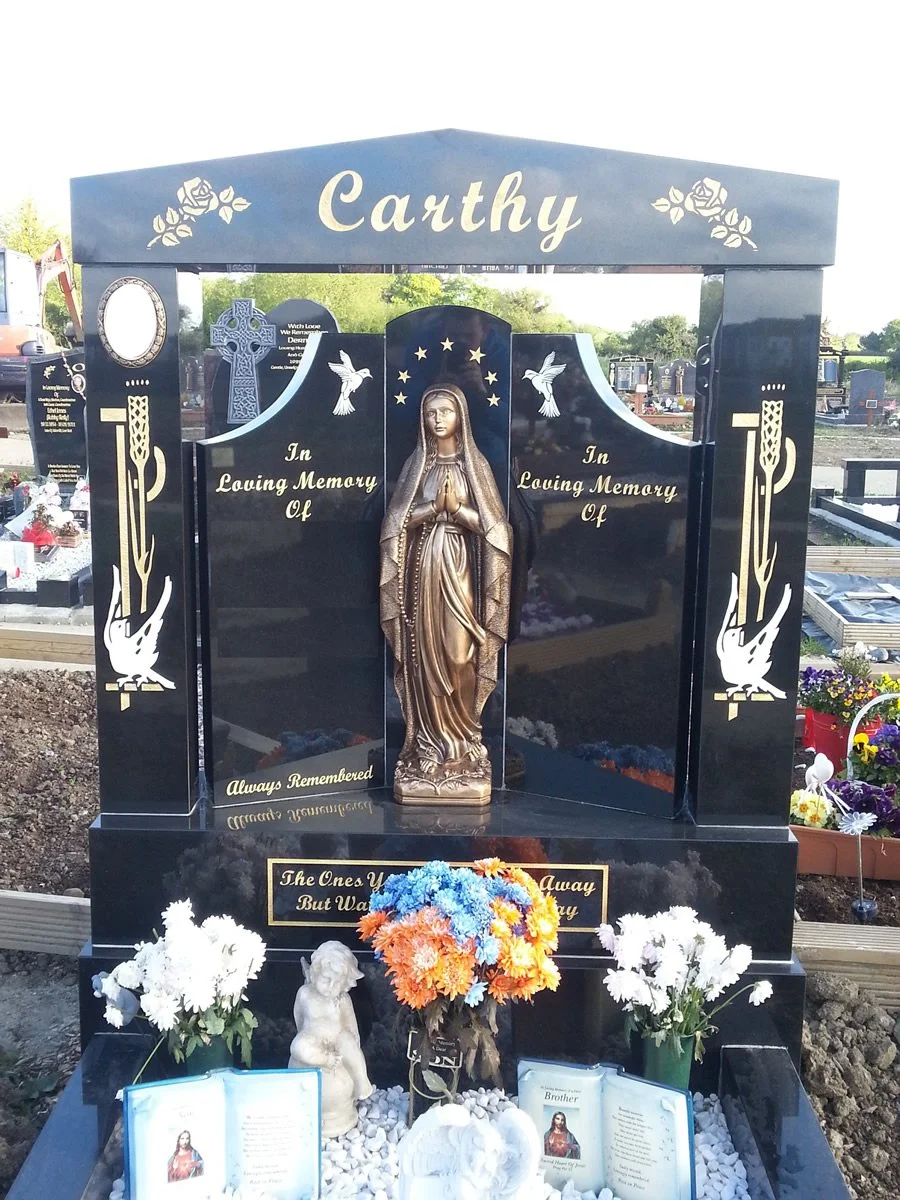 A black granite gravestone with a gold statue of the Virgin Mary in prayer in the center. The top has 'Carthy' written with floral designs. On either side of the statue, there are inscribed words 'In Loving Memory Of.' The gravestone is decorated with flowers, including a bouquet of orange and blue flowers and white carnations, and smaller flower arrangements. Religious symbols, such as doves, are also inscribed on the gravestone.