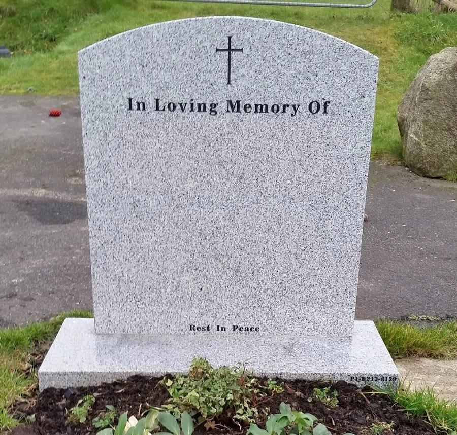 Gray granite headstone with a cross at the top, inscribed with 'In Loving Memory Of' and 'Rest In Peace' at the bottom, in a grassy cemetery area.
