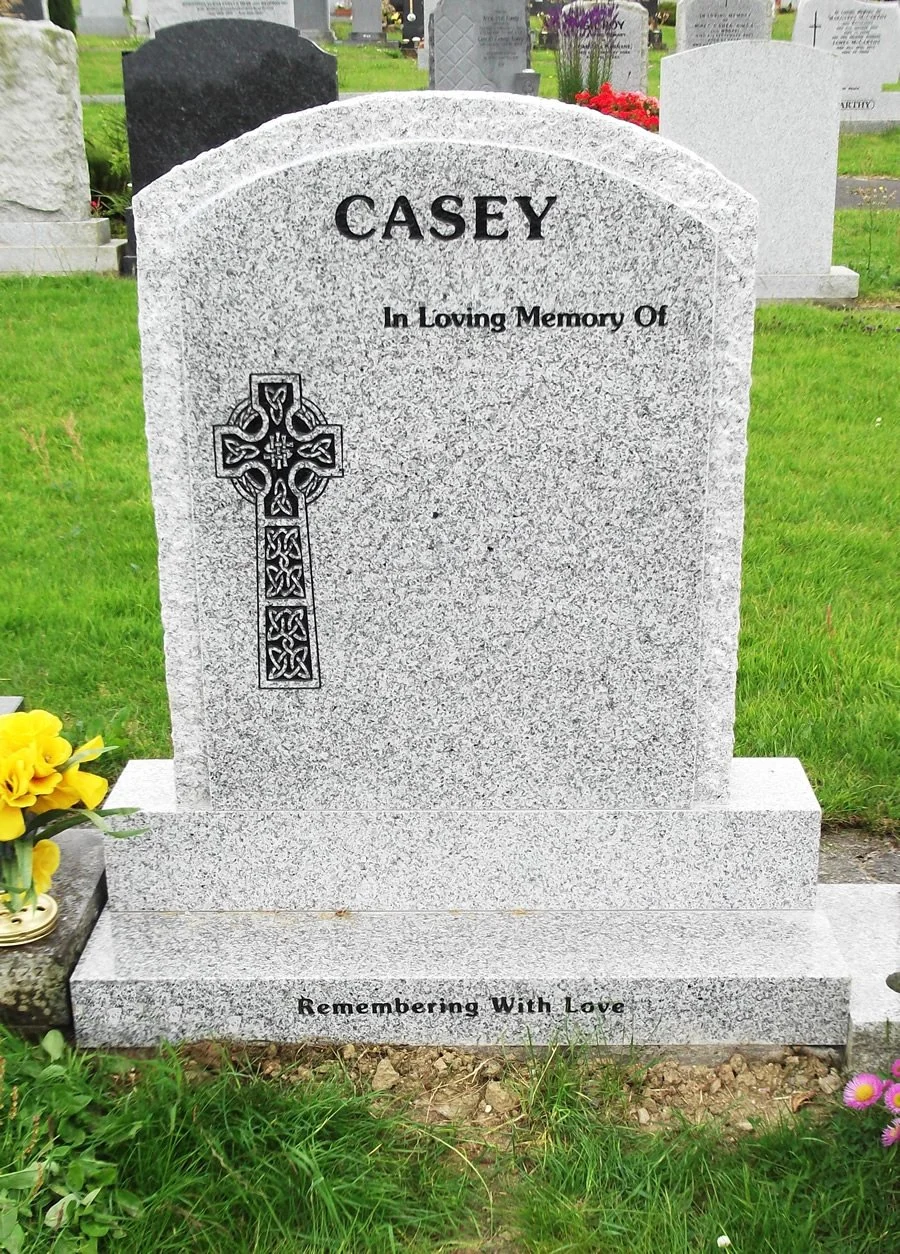 Gray granite headstone in a cemetery with the name 'Casey' at the top, a Celtic cross on the left side, and the inscription 'In Loving Memory Of' in the center. At the bottom, it says 'Remembering With Love.' The headstone is surrounded by green grass and some flowers.