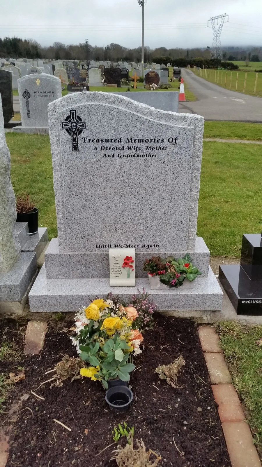 Gravestone in a cemetery with flowers and a small card, reading 'Treasure Memories Of A Devoted Wife, Mother, And Grandmother. Until We Meet Again.'