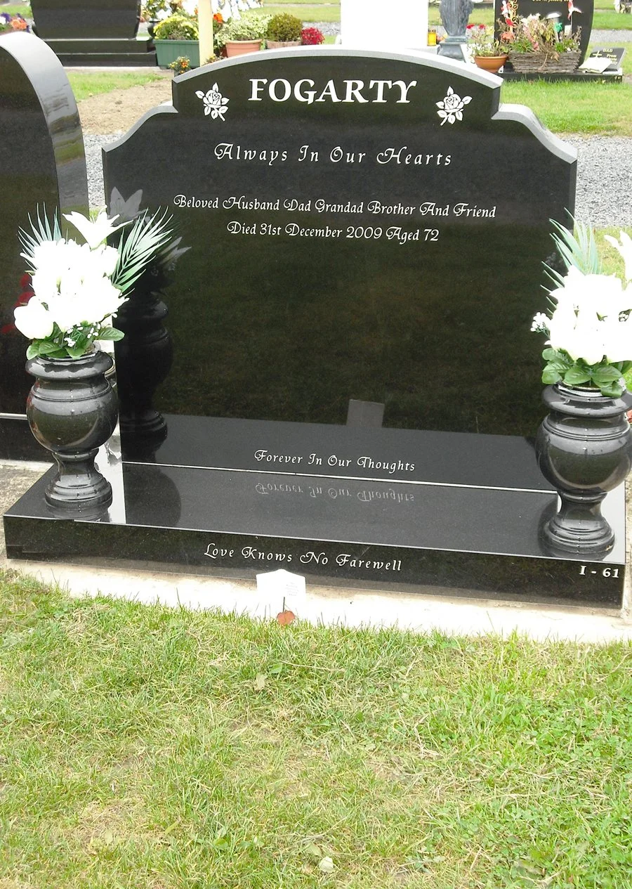 Black granite headstone for Fogarty family with engraved white text; floral decorations and urns with white flowers on each side; grass in foreground and surrounding flowers and monuments in background.