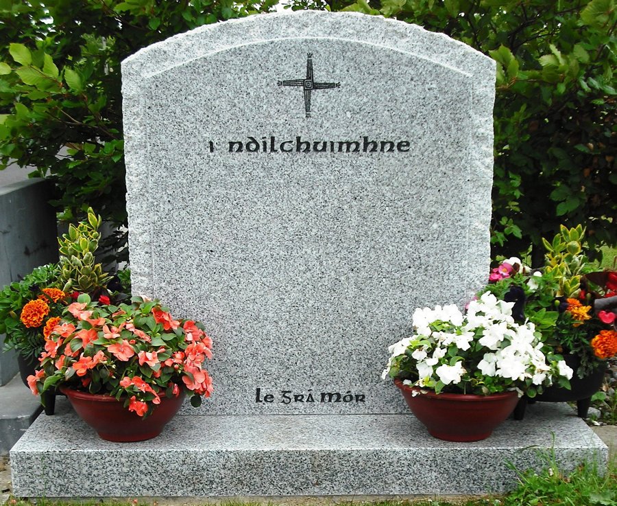 A granite gravestone with a cross at the top, surrounded by flower pots with colorful flowers, and green shrubbery in the background.