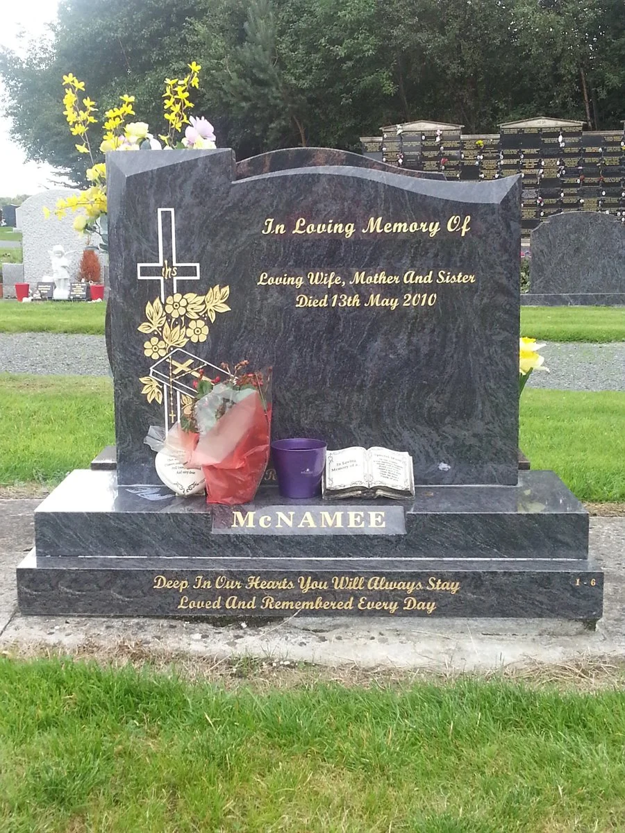 A black granite gravestone with gold and white inscriptions, flowers, a small cross, a photo, and a book placed on it, in a cemetery with other gravestones and greenery in the background.