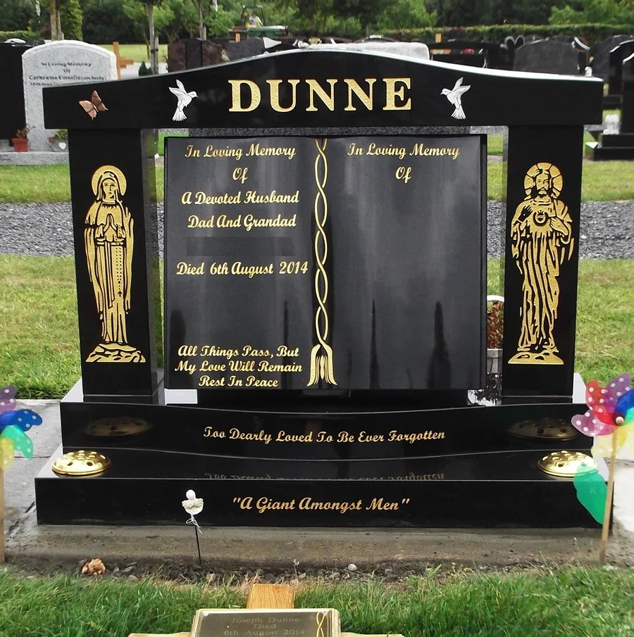 Black granite headstone with gold inscription and angel illustrations, dedicated to Dunne, a husband, dad, and granddad who died on August 6, 2014, situated in a cemetery with other headstones in the background and colorful pinwheels at the base.