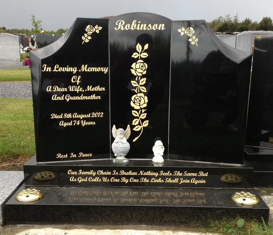 A black granite tombstone with gold engraving, dedicated to Robinson, in memory of a wife, mother, and grandmother who died on August 8, 2012, at age 74. The tombstone features gold roses and a vertical stem of roses with leaves. There are two angel figurines at the base and engraved text at the bottom about family bonds.