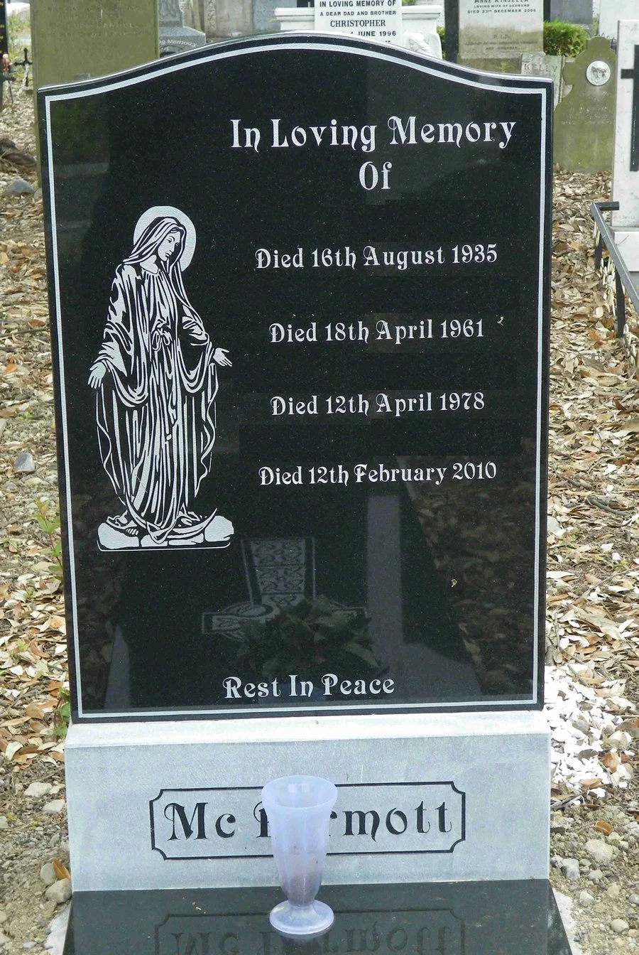 A gravesite memorial headstone in a cemetery. It has a black polished surface with white engraved text and an illustration of the Virgin Mary. The gravestone reads: "In Loving Memory of" with death dates of four individuals, ending with "Died 12th February 2010." The bottom has the phrase "Rest In Peace" and a small vase in front.