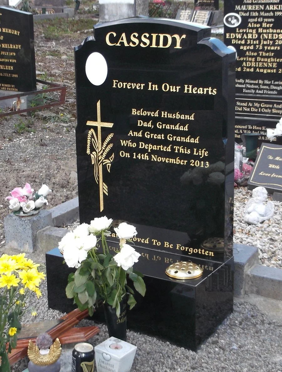 A black tombstone with the name 'CASSIDY' at the top, inscribed with a message honoring a loved one who passed away on November 14, 2013. The tombstone has a gold cross with wheat and ribbon design, and flowers are placed in front. There are additional memorial items and flowers around the grave.