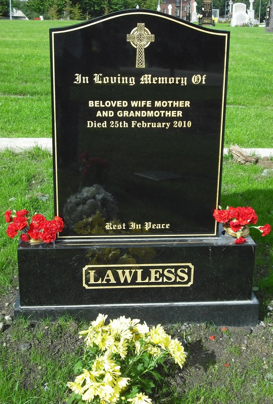 Gravestone with a Celtic cross at the top, inscribed with a memorial message for a beloved wife, mother, and grandmother who died on February 25, 2010. The gravestone is black with gold lettering, and there are red flowers on either side and yellow flowers at the base. The word 'LAWLESS' is engraved at the bottom.