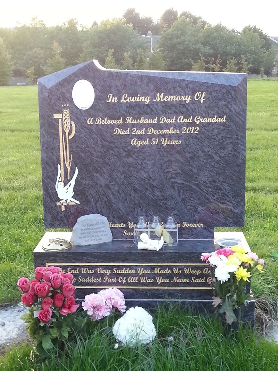 Gravestone with an illustration of a white angel holding a cross, surrounded by pink, white, and yellow flowers, and candles. The inscription honors a husband and grandfather who died in December 2012 at age 51.