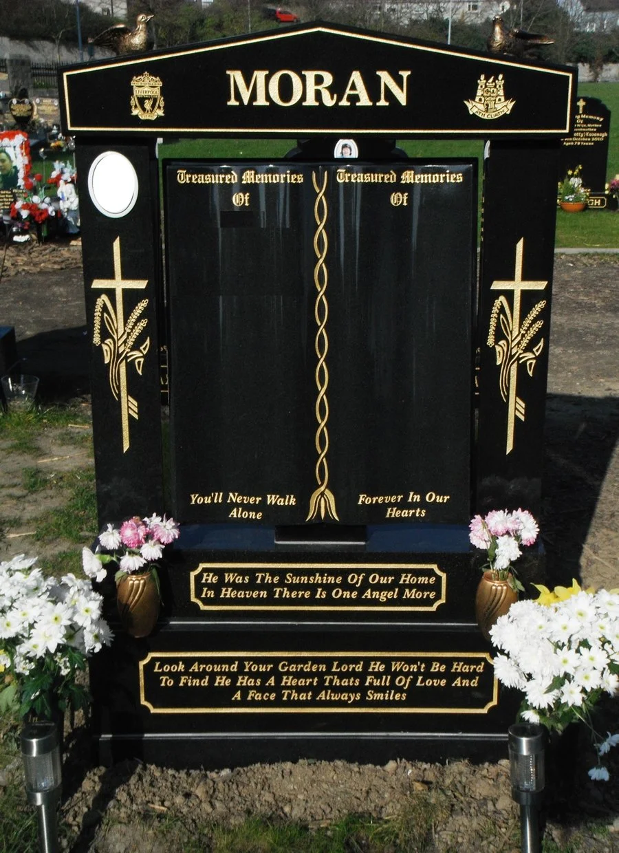 A black headstone with gold accents in a cemetery. The headstone has the name "Moran" at the top, religious symbols on the sides, and floral decorations at the base. It has inscriptions including "Treasured Memories of," "You'll Never Walk Alone," "Forever In Our Hearts," and a poem about an angel in heaven.