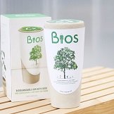 Two white biodegradable plant pots and a white travel mug with green tree artwork on a wooden surface.