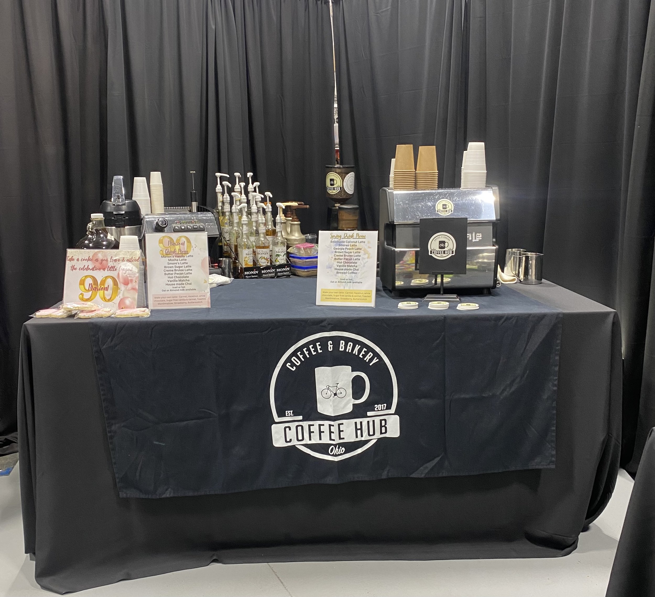 A black tablecloth with a white circular logo reads 'Coffee Hub Ohio.' Behind the table, there are black curtains, and on the table, there are cups, a coffee machine, and a sign listing various coffee drinks.
