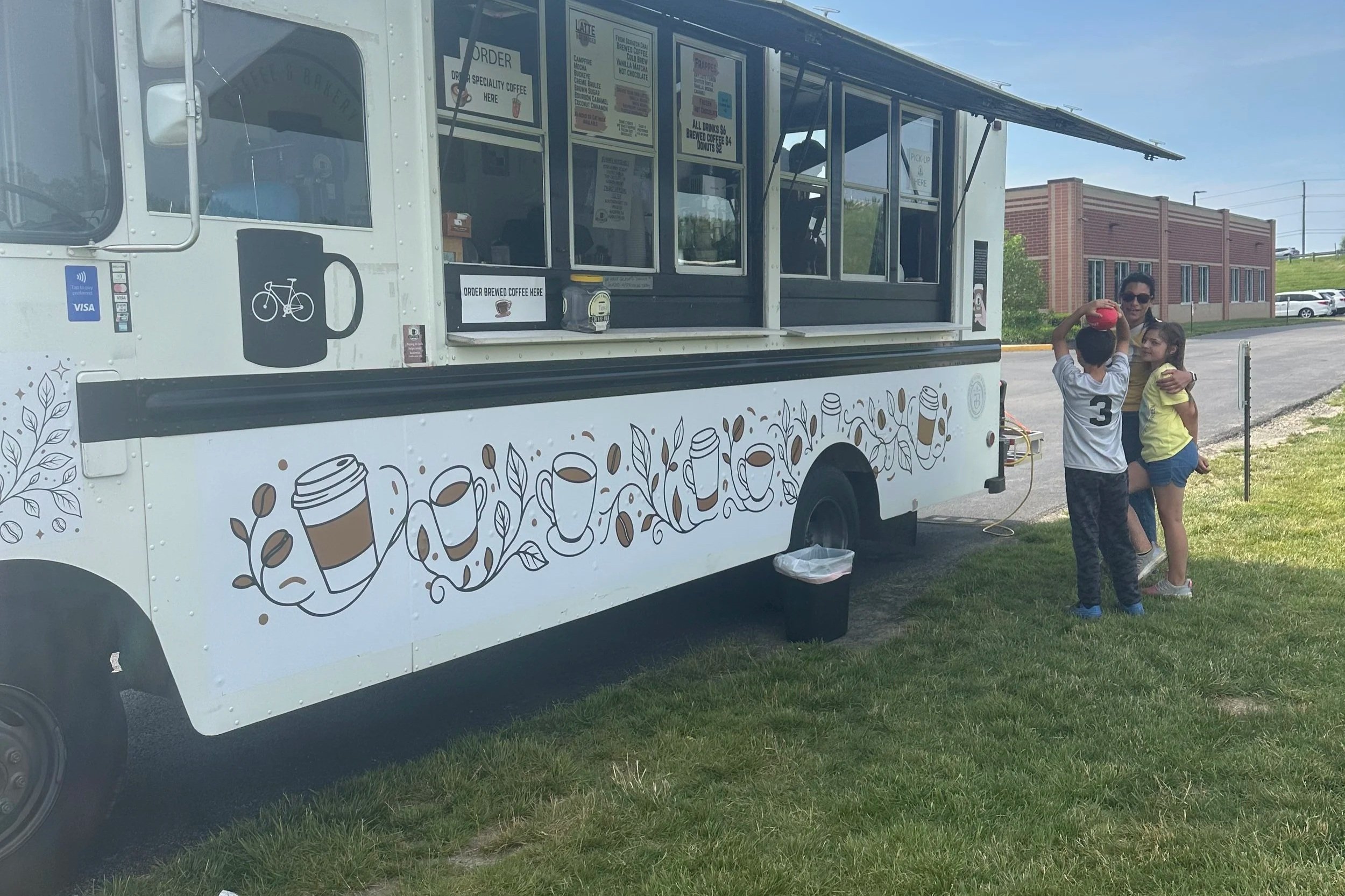 A food truck with coffee-themed graphics and signs, parked on a grassy area. Two children and an adult are standing nearby, with some of the children holding a red object. The scene is outdoors on a clear day.