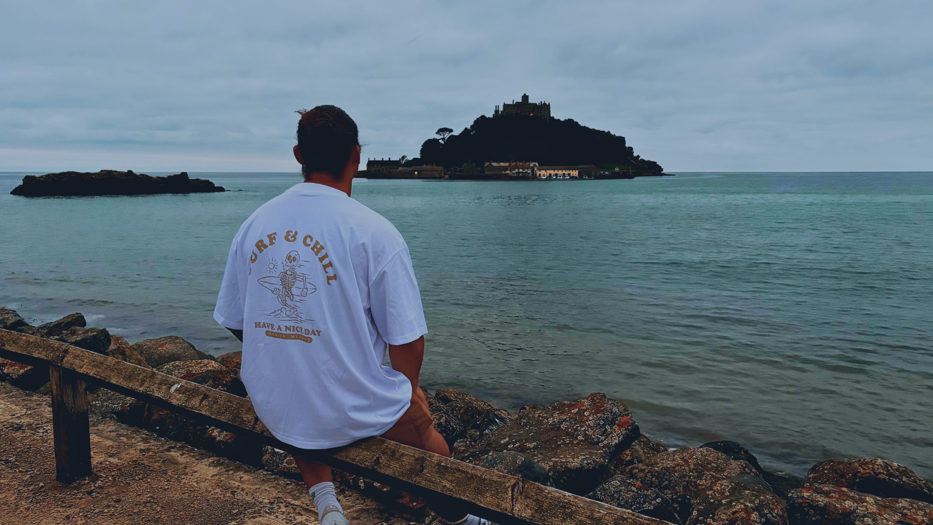A man sat looking at St Michael's Mount wearing a top by Cornish Mermaid that says "Surf & Chill"