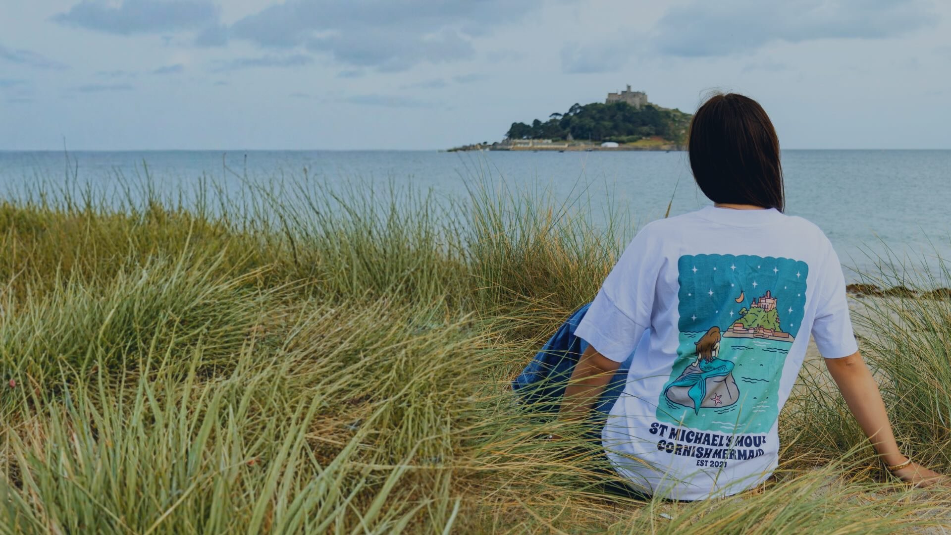 A girl sat looking at St Michael's Mount wearing a graphic Cornish Mermaid T Shirt