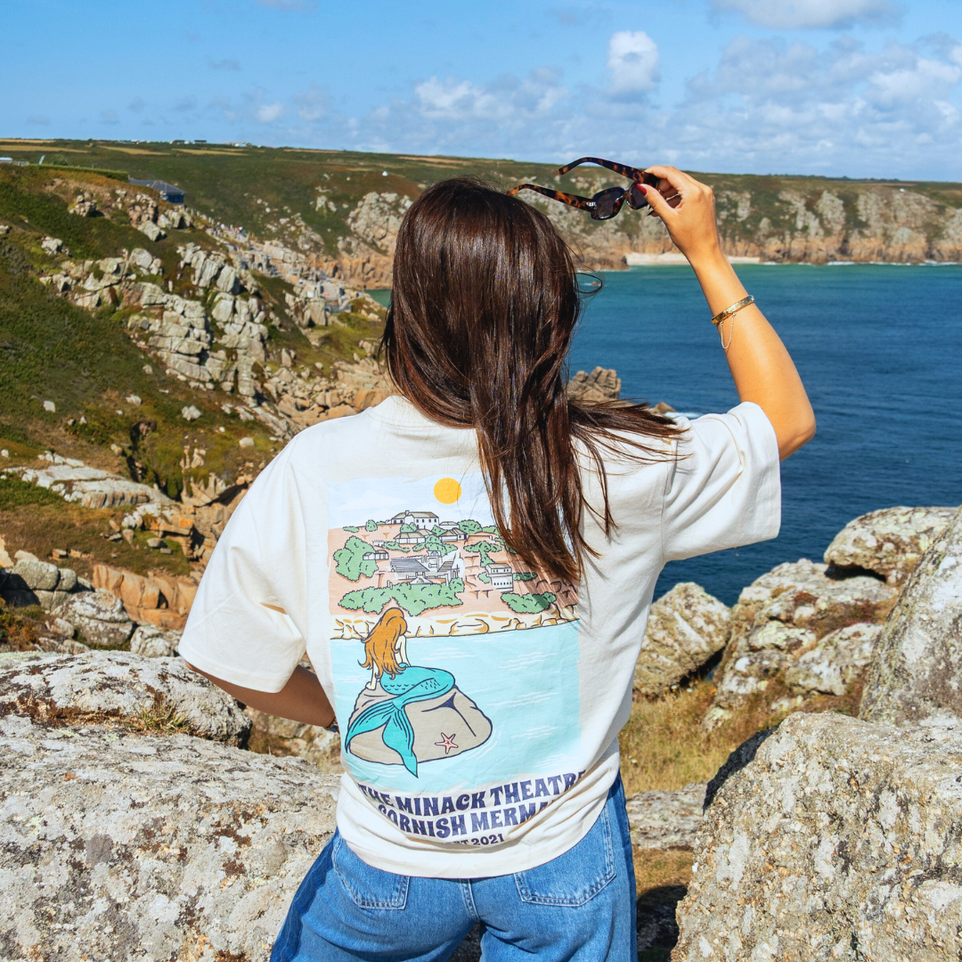 A girl facing off a cliff wearing a Tee Shirt of the Minnack Theatre in Cornwall