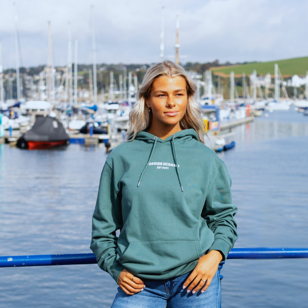 Young woman standing by a marina with boats and sailboats in the background, wearing a teal hoodie and blue jeans.