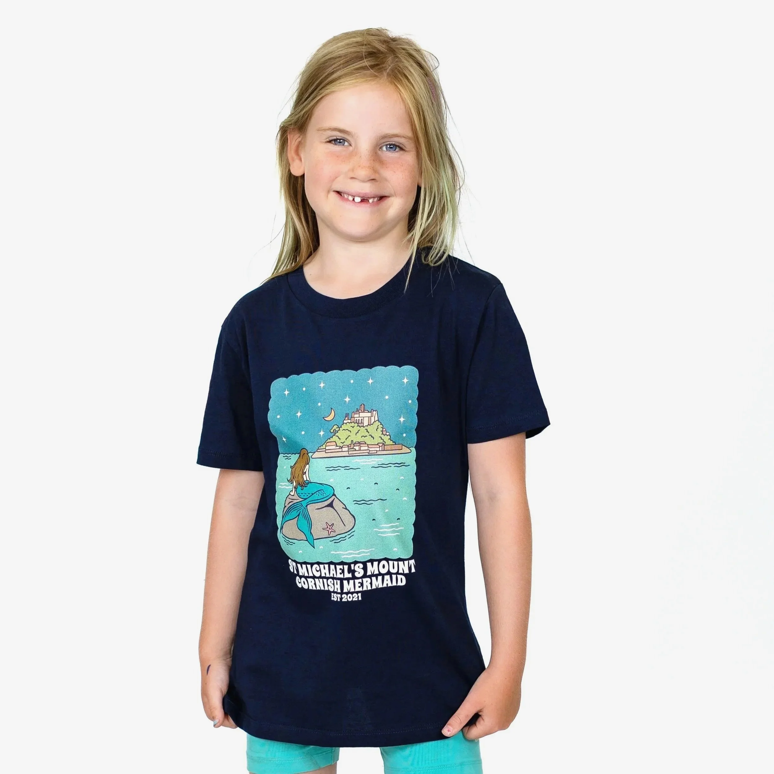 Young girl smiling, wearing a dark blue T-shirt with a graphic of a mermaid sitting on a rock and an island with a castle in the background. The T-shirt has text reading "St. Michael's Mount, Cornish Mermaid, EST 2021."