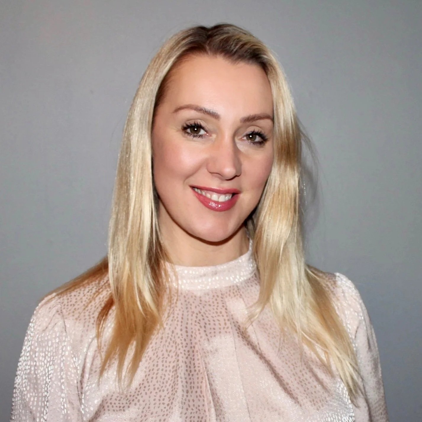 Portrait of a smiling woman with long, blonde hair, wearing a light, silky blouse, standing indoors against a light gray background