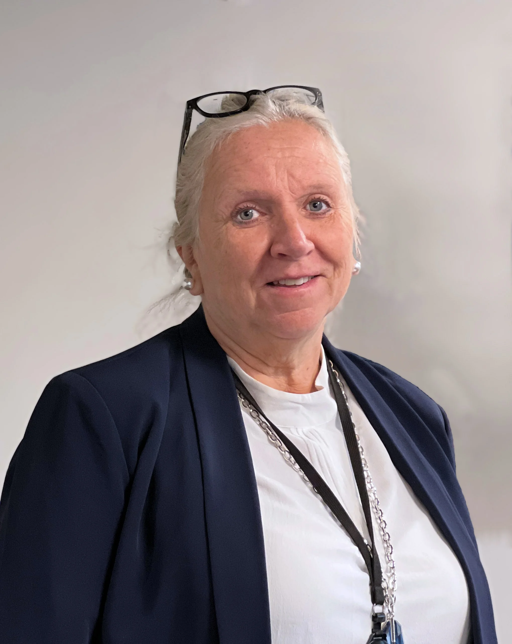 Portrait of a woman with light hair tied back, wearing glasses on top of her head, a white blouse, and a dark blazer, smiling at the camera against a light gray background